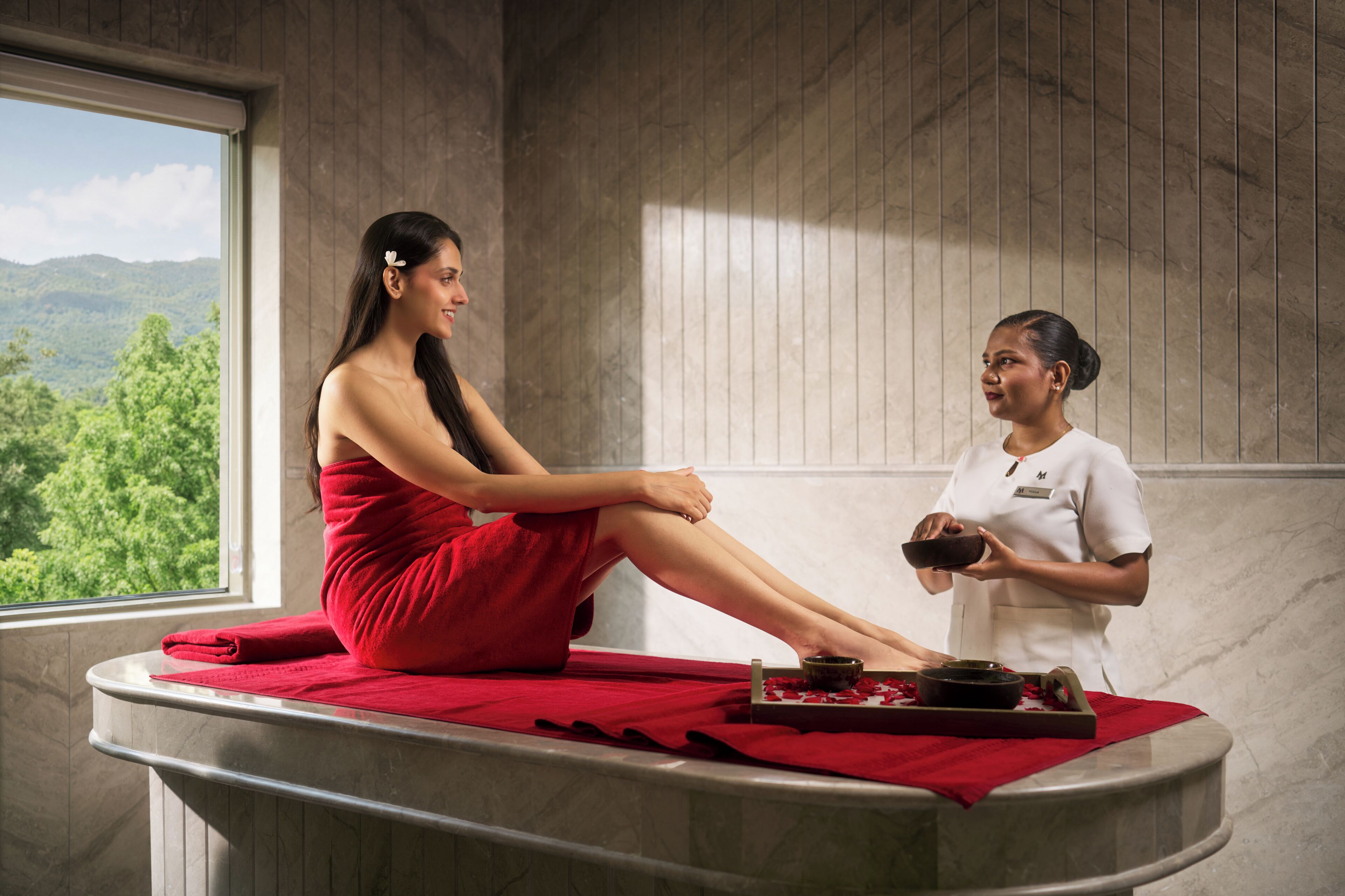 A spa setting with a person seated on a red towel, another person holding a bowl, and a tray with items on a marble counter.