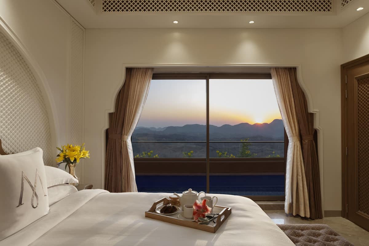 A hotel room with a large bed, a tray with breakfast items, and a view of mountains at sunset.