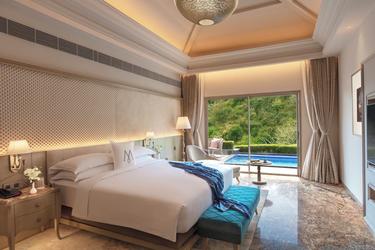 A spacious bedroom with a large bed, a sitting area with a view of a pool, and a balcony with curtains.