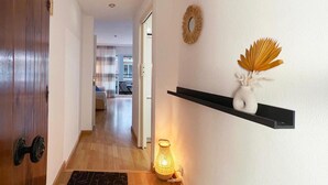 Un couloir avec une porte en bois, un paillasson noir, une étagère fixée au mur avec un vase et des feuilles décoratives, et un lampadaire allumé.