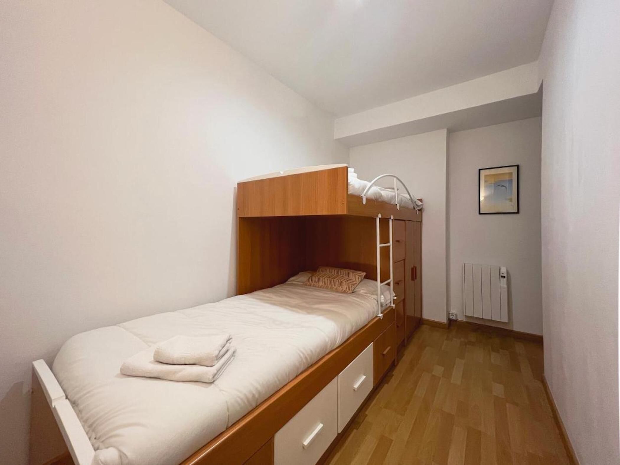 Une chambre avec un lit superposé, une tête de lit en bois, un matelas blanc et un lit simple avec une couverture blanche. Un radiateur blanc est fixé au mur, et un tableau encadré est accroché au-dessus.
