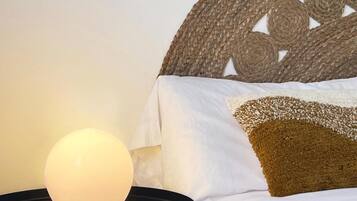 Una cama con ropa de cama blanca y marrón, una mesa redonda con una lámpara y un cabecero tejido.