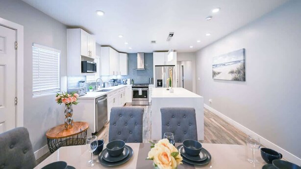 A modern kitchen with white cabinets, a central island, and a dining area with grey upholstered chairs and a table set for four. - Stylish Central San Jose 3B2B w/crib Free Parking (San Jose)