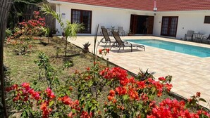 Un espace aménagé au bord de la piscine, avec une terrasse carrelée, des chaises longues et un petit jardin agrémenté de fleurs rouges.