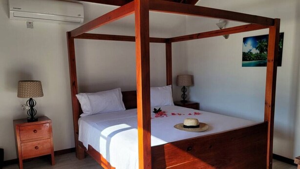 Un lit à baldaquin avec un cadre en bois, du linge de lit blanc et un chapeau posé sur le lit. Il y a une table de chevet avec une lampe et un tableau accroché au mur.