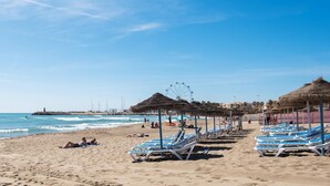 A beach with rows of sun loungers and thatched umbrellas, a clear blue sky, and a distant view of a Ferris wheel. - Carihuela Beach Sun Private apartment (TORREMOLINOS)