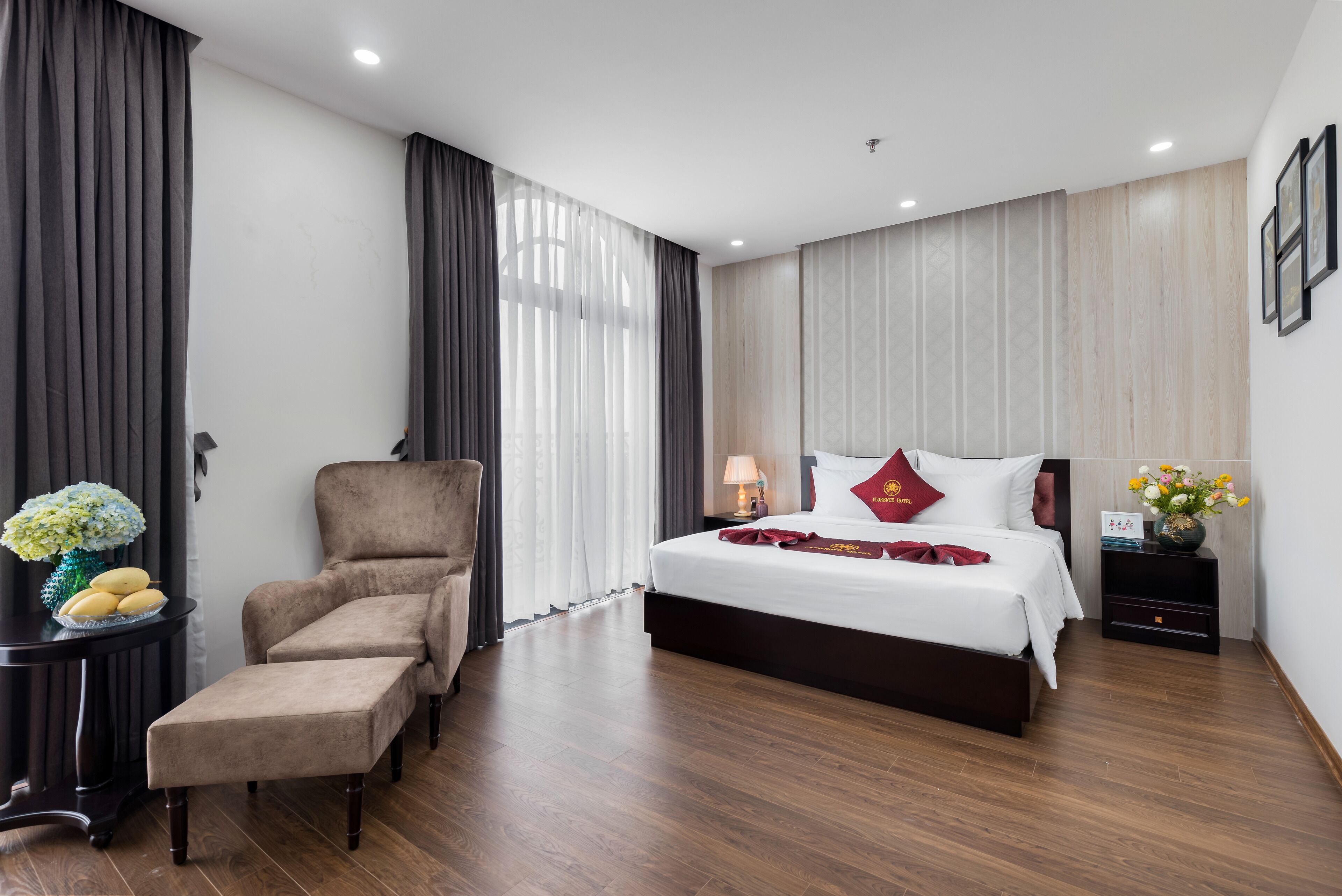 Una habitación de hotel con una cama grande, una silla, una mesita con un jarrón y una mesita de noche.