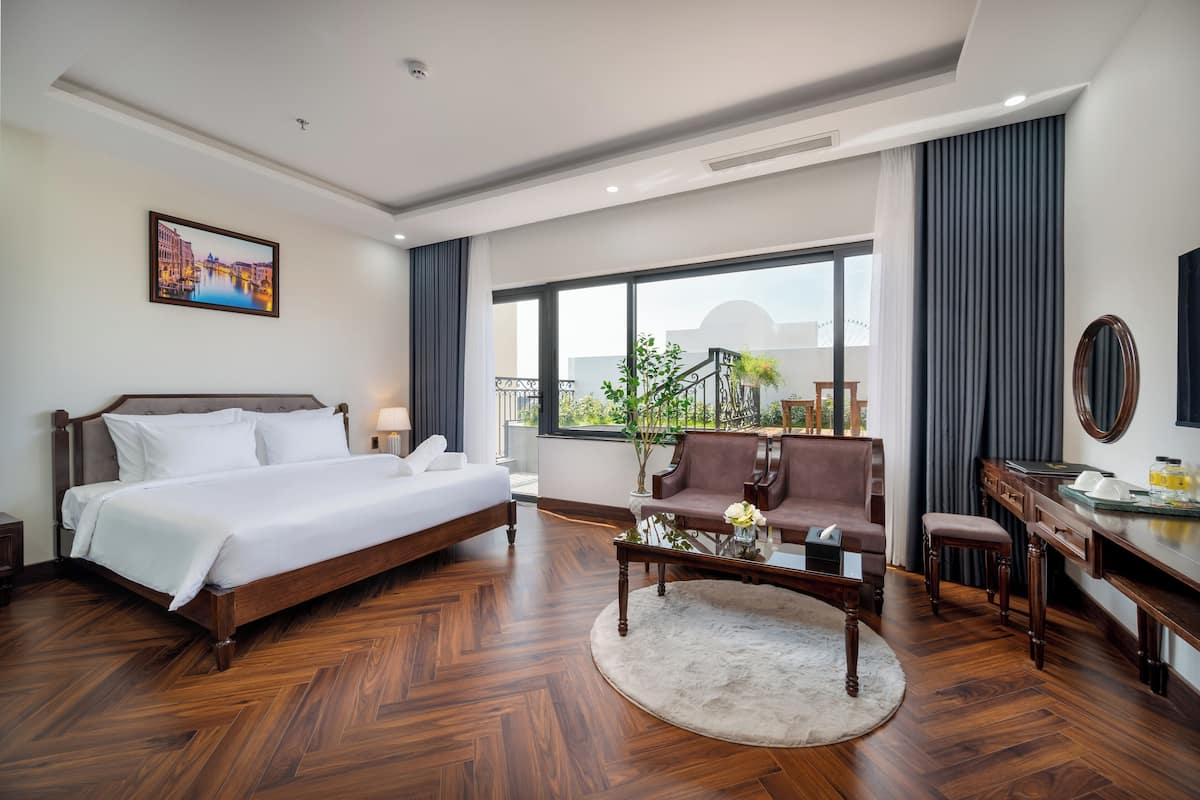 A hotel room with a large bed, a sitting area with a sofa and armchair, a desk with a chair, and a large window offering a view of the outdoors.