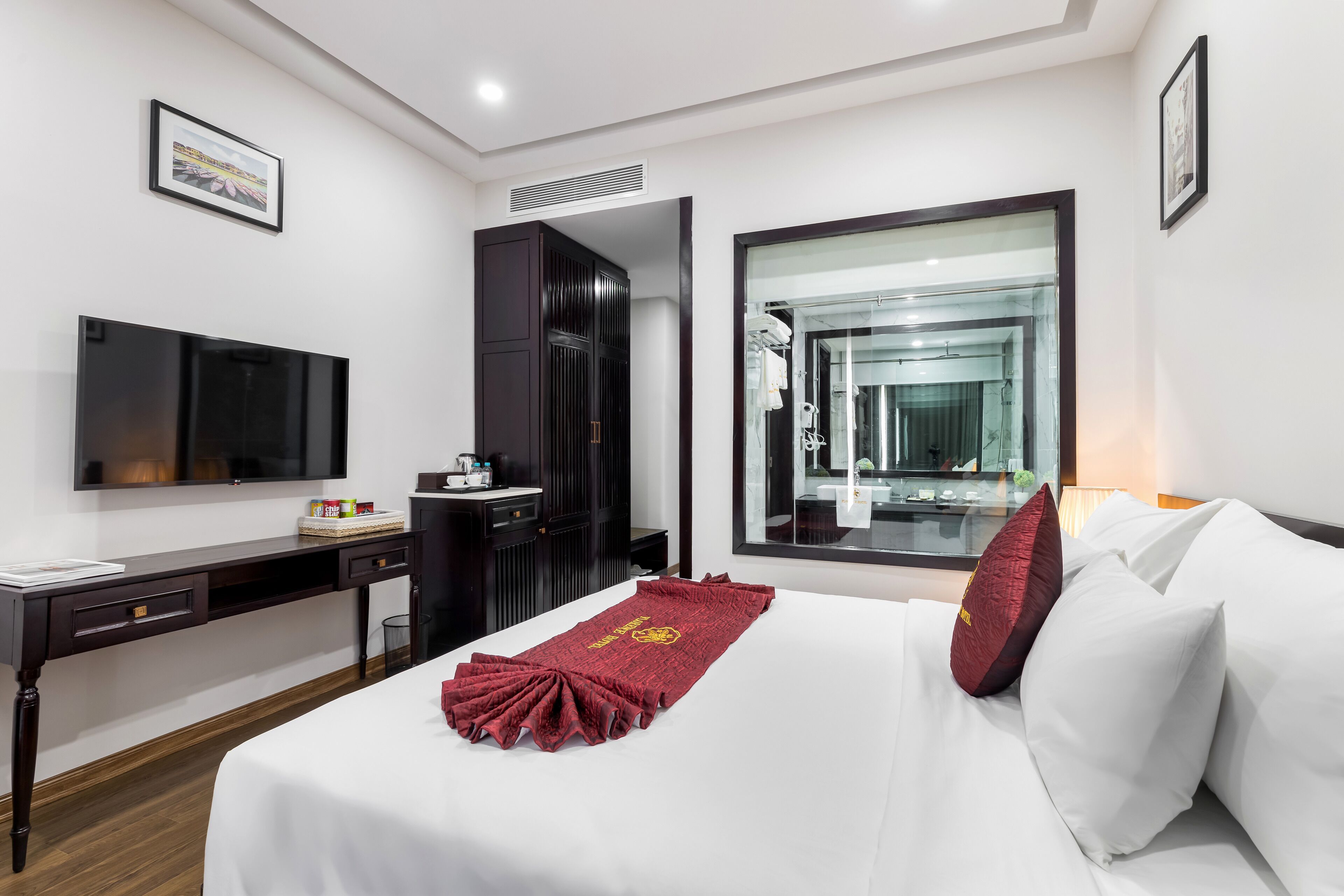 A hotel room with a bed, a desk, a television, a wardrobe, and a bathroom visible through a glass door.