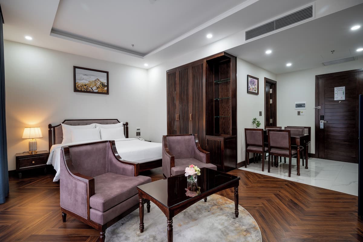 A hotel room with a bed, two armchairs, a coffee table, a desk with chairs, and a wooden wardrobe.