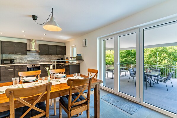 A modern kitchen with a dining area, large sliding glass doors to an outdoor patio, and a view of greenery.