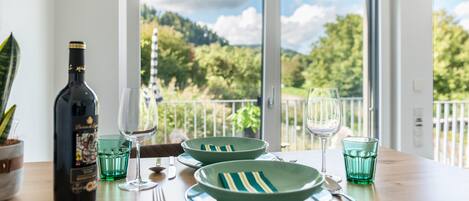 A dining table with plates, cutlery, and a bottle of wine, set against a backdrop of a balcony with a view of greenery.