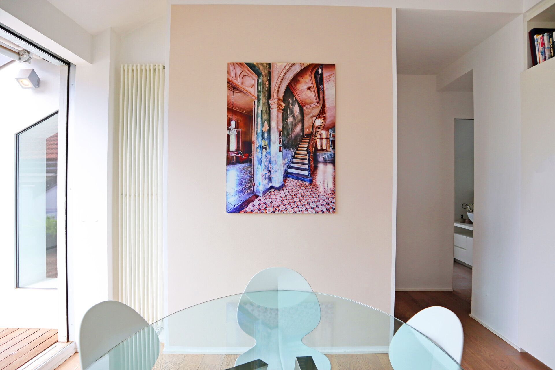A modern interior with a glass table, a large painting of an architectural corridor, and a staircase.