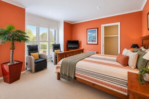 A bedroom with a bed, a television, a chair, a sofa, a plant, and a window.