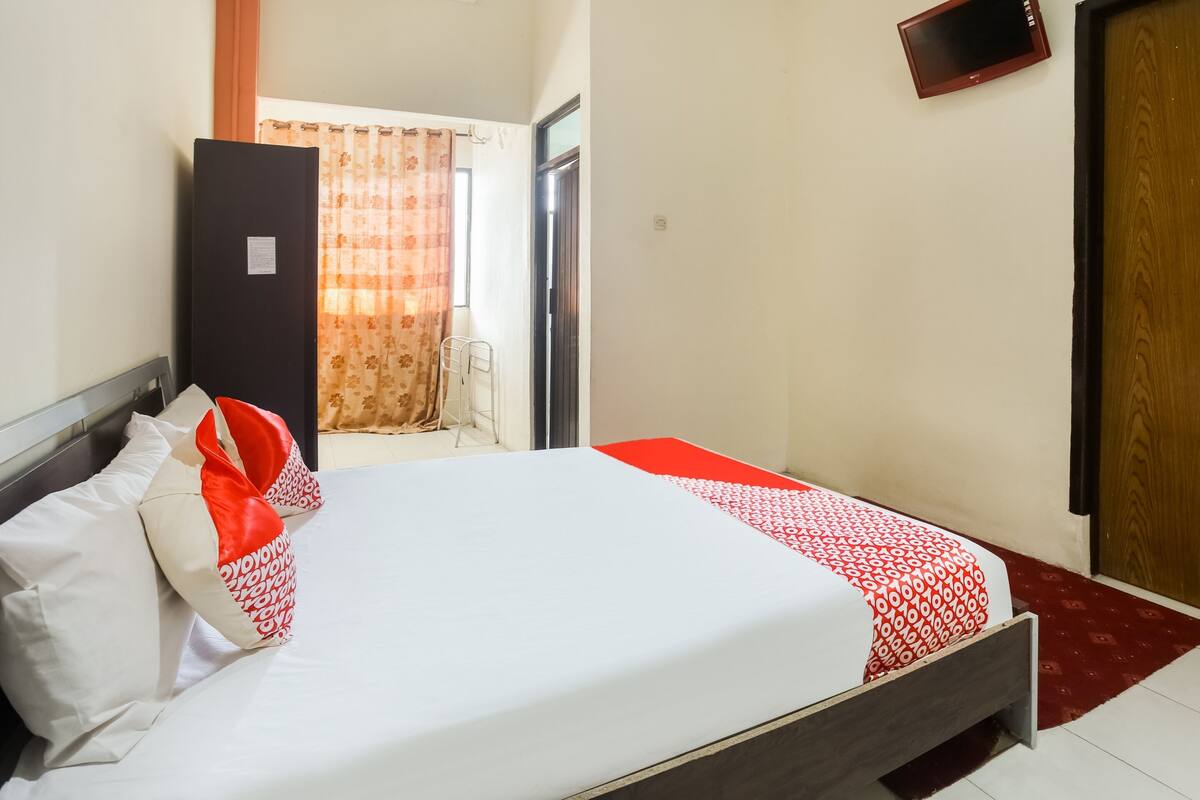 Una cama bien hecha con una colcha blanca y roja estampada, un cabecero de madera y un almohadón rojo y blanco.