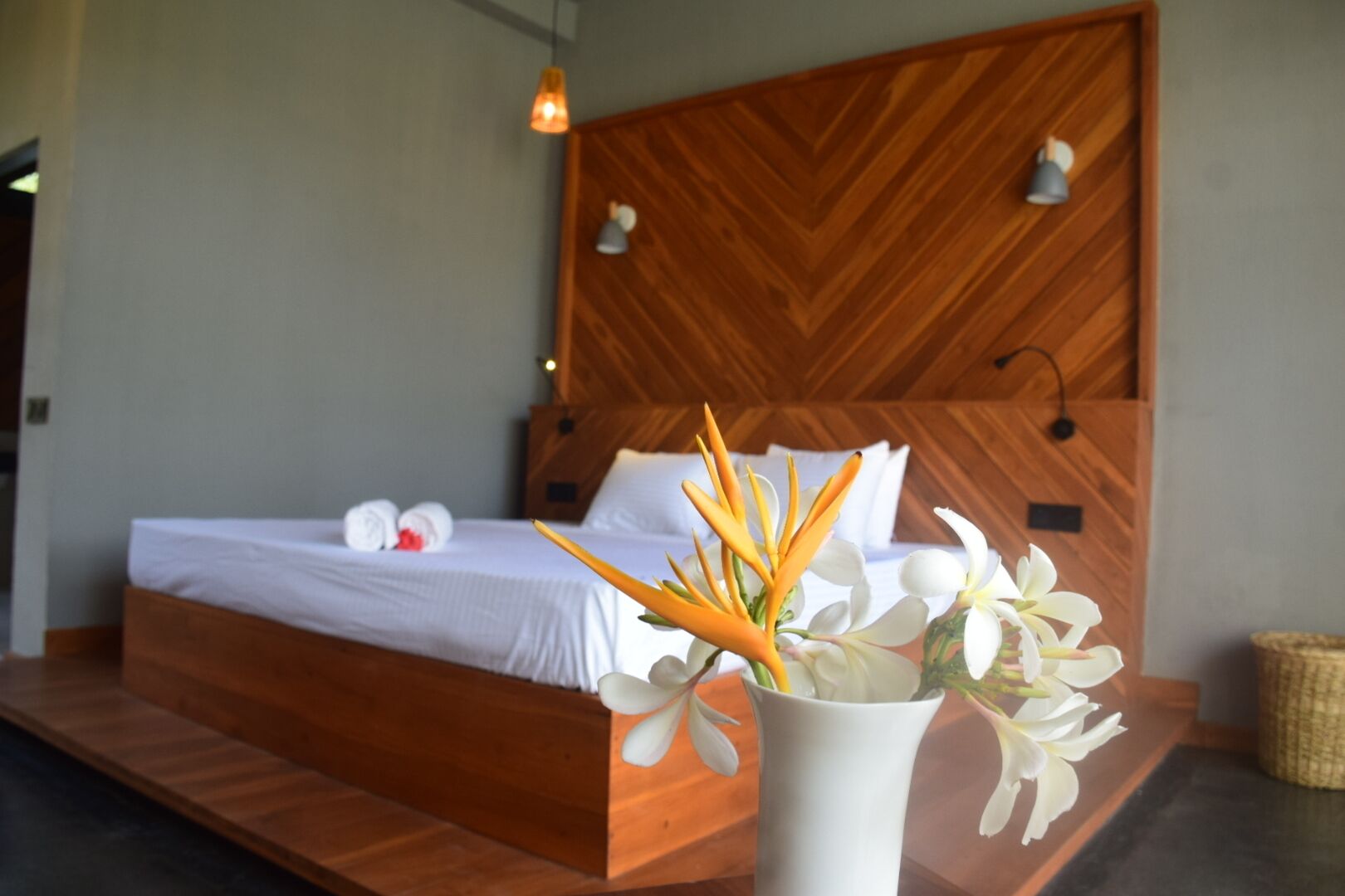 Una cama bien hecha con sábanas blancas y un cabecero de madera, un jarrón con flores blancas y una pequeña cesta tejida en el suelo.