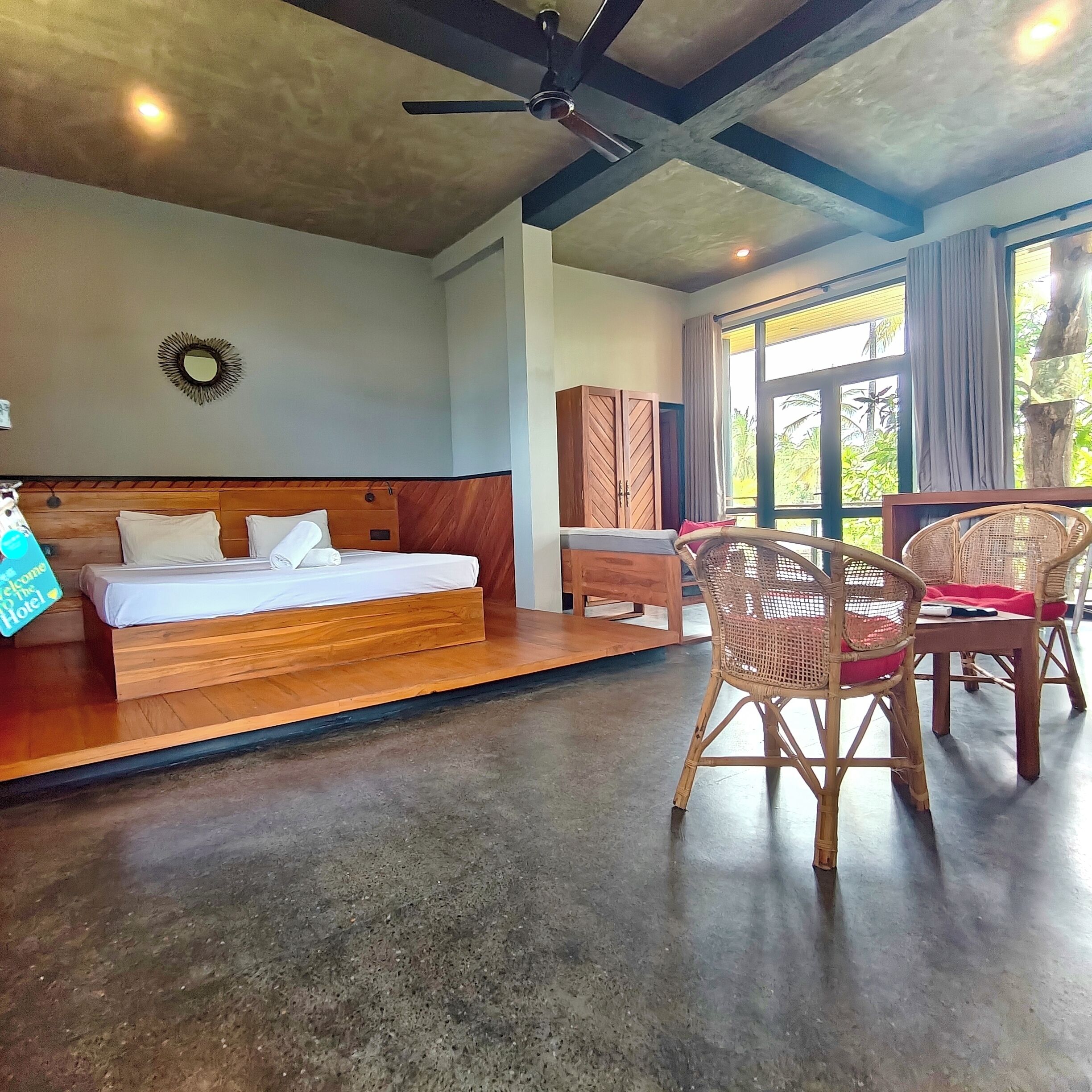 A room with a bed, a desk, wicker chairs, a ceiling fan, and a large window.