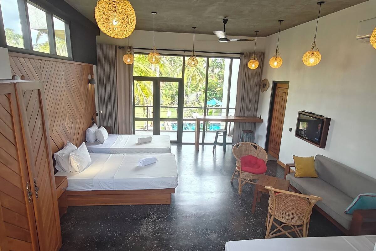 A hotel room with two beds, a sofa, a TV, and a balcony with a view of greenery.
