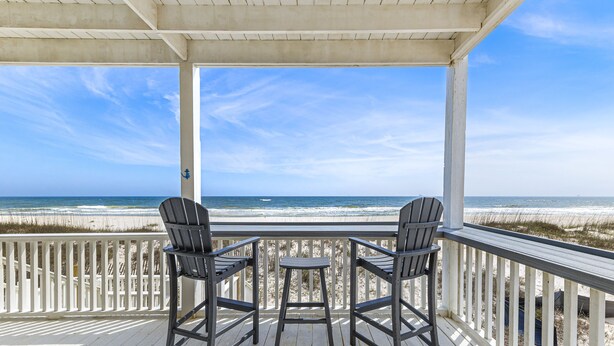 Una terraza cubierta con dos sillas Adirondack y una pequeña mesa, con vista a una playa con dunas de arena y el océano.