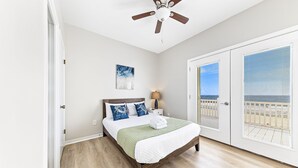 Un dormitorio con cama de madera, ventilador de techo, mesita de noche con lámpara y vista a la playa a través de una puerta corrediza.