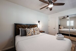 Ein Schlafzimmer mit hölzernem Kopfteil, einem Bett mit weißer Bettwäsche, einem Deckenventilator, einem Nachttisch und einem Spiegel, der den Raum reflektiert.