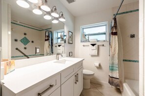 Ein Badezimmer mit weißem Waschbecken, Spiegel und Toilette. Ein Fenster bietet Blick aufs Meer, und ein Duschvorhang zeigt ein Strandmotiv.