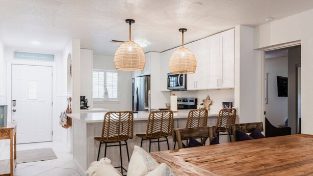 Una cocina moderna con armarios blancos, una mesa de comedor de madera, sillas de mimbre y una lámpara colgante.