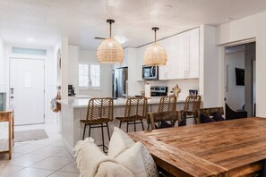Eine moderne Küche mit weißen Schränken, einem Holztisch, Korbstühlen und einer Pendelleuchte.