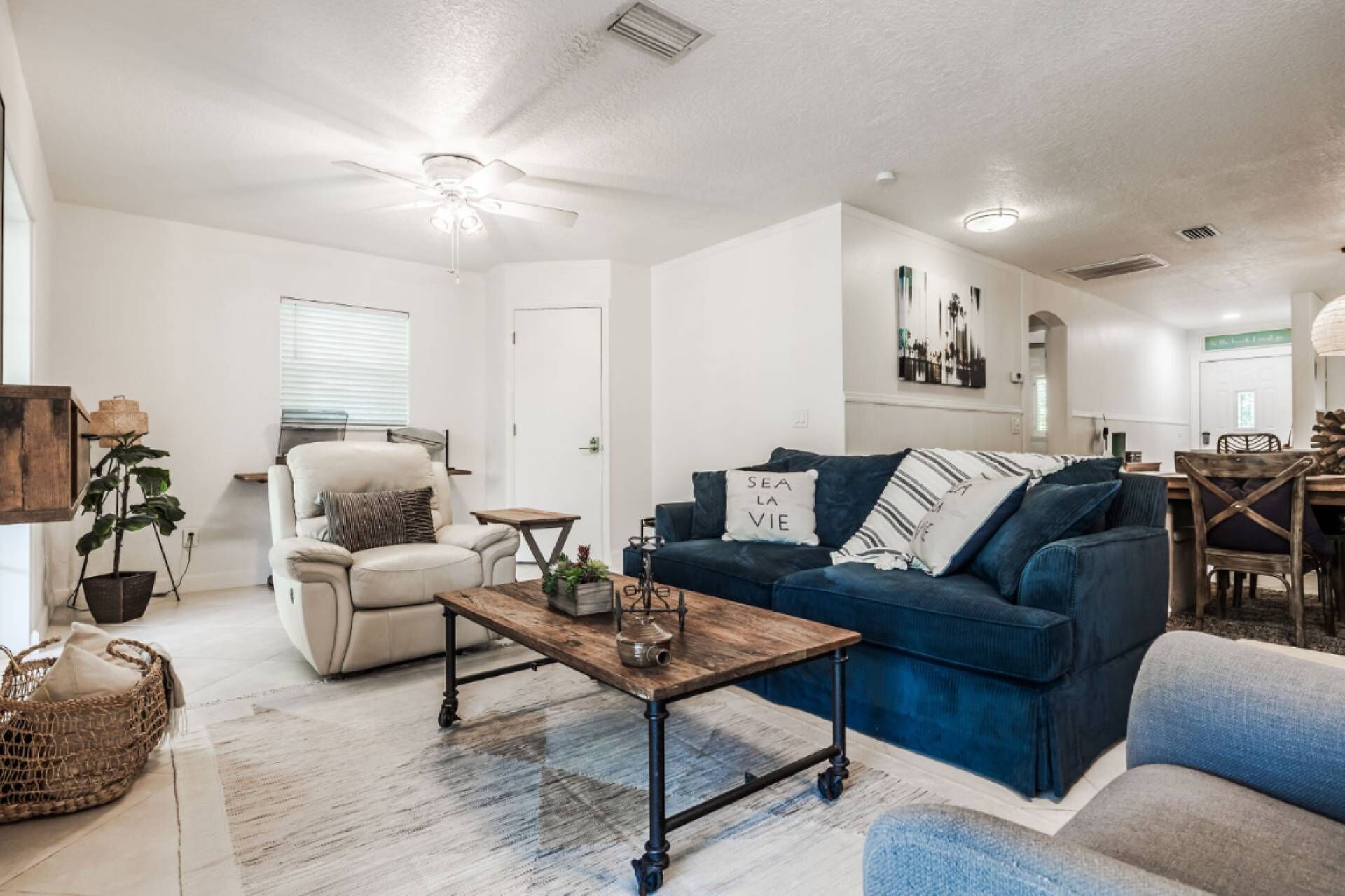 A spacious living room with a white ceiling, a ceiling fan, a blue sofa, a white armchair, and a wooden coffee table.