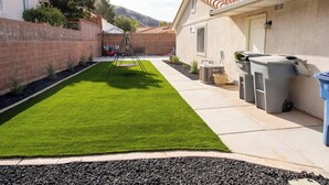 A backyard with artificial grass, a swing set, a patio area with outdoor furniture, and a house with a tiled roof. - Private home near Zion & state parks, BBQ, cozy stay for family & friends in scenic red rock views! (La Verkin)