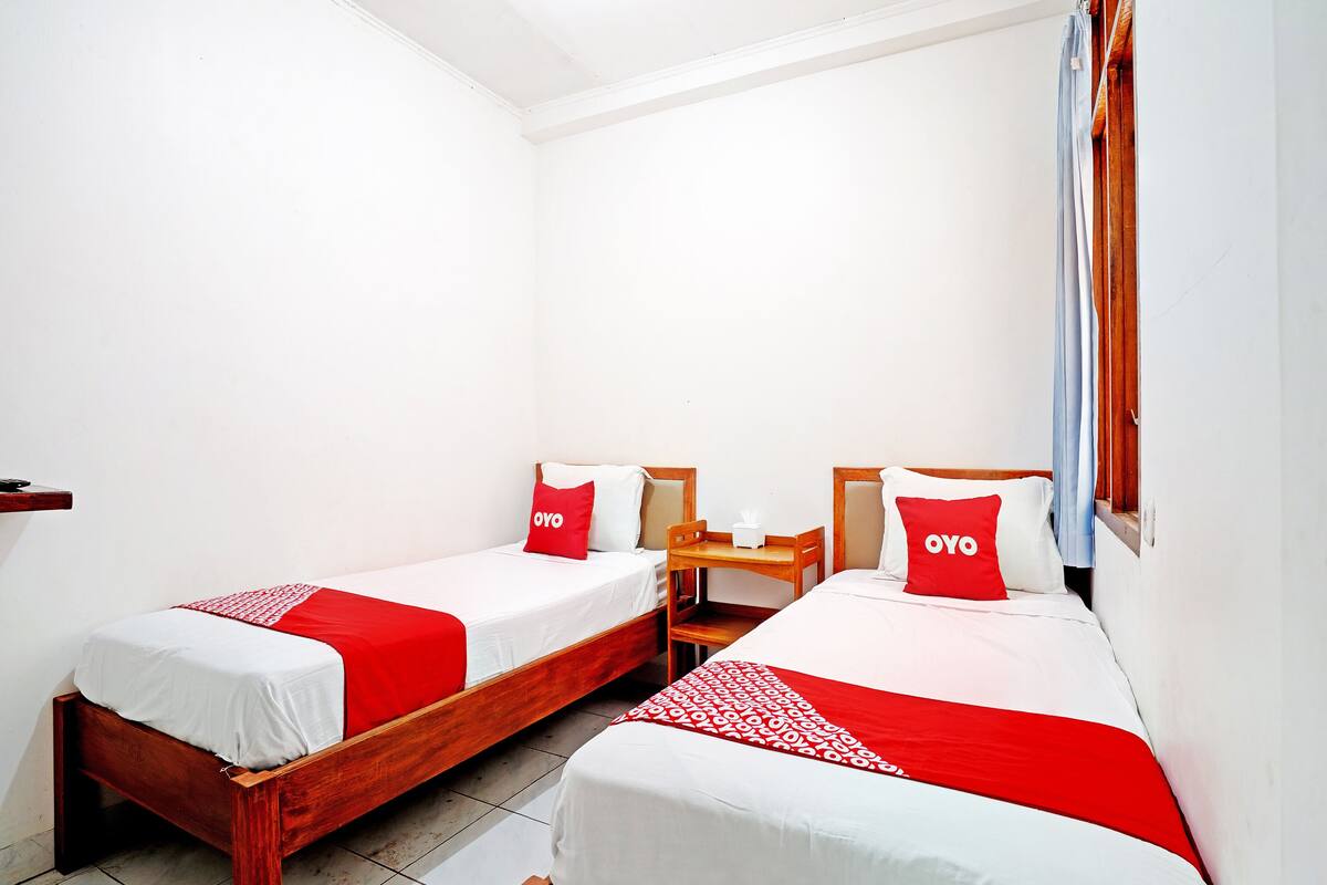 Une chambre d’hôtel avec deux lits simples, chacun doté d’un cadre en bois et d’une literie rouge et blanche. Une petite table en bois se trouve entre les lits, et une boîte de mouchoirs y est posée.