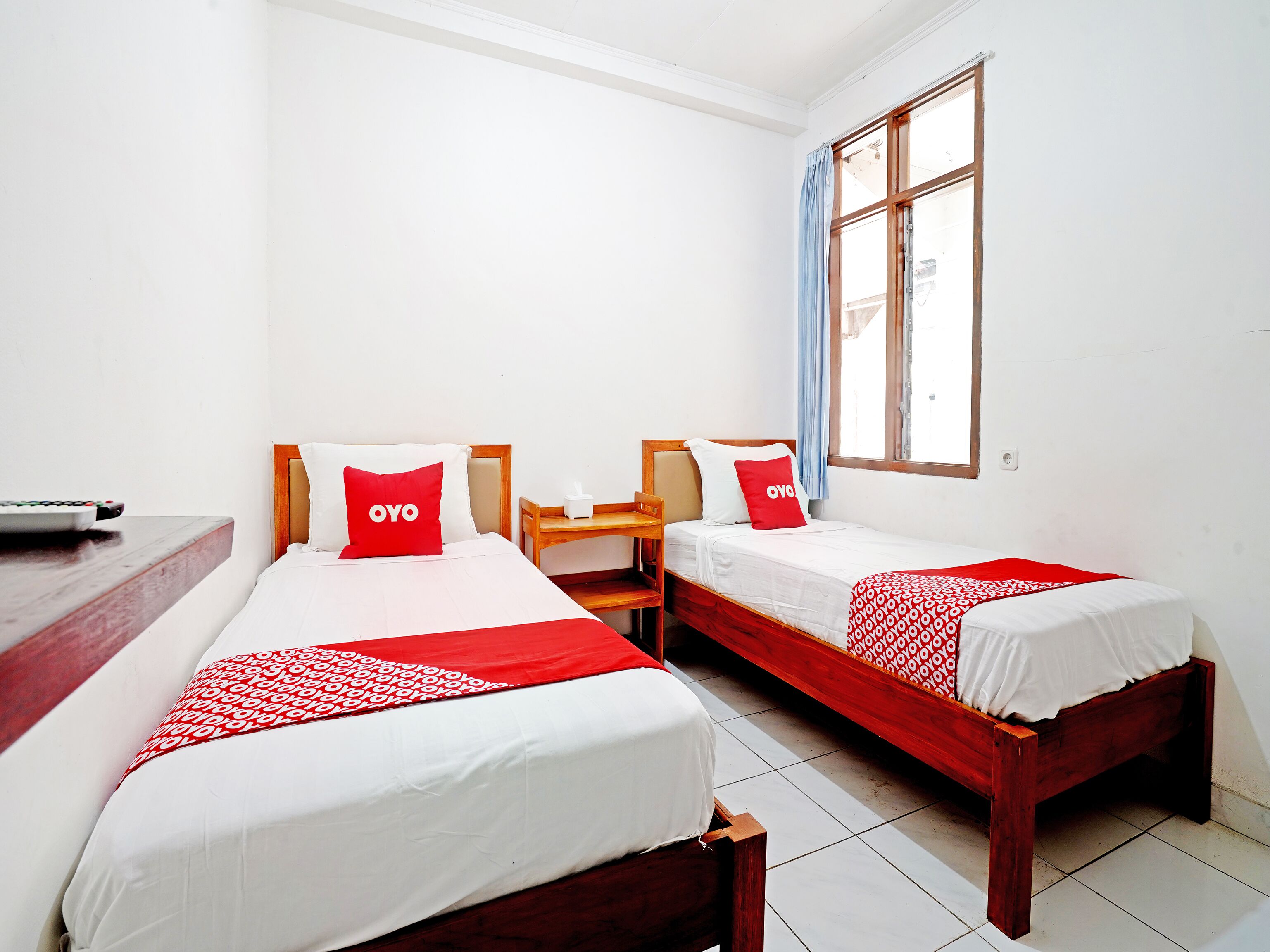 A hotel room with two single beds, each with a red and white bedspread and matching pillows. There is a wooden nightstand with a lamp and a tissue box between the beds. A wooden desk with a remote control is visible on the left side of the image.