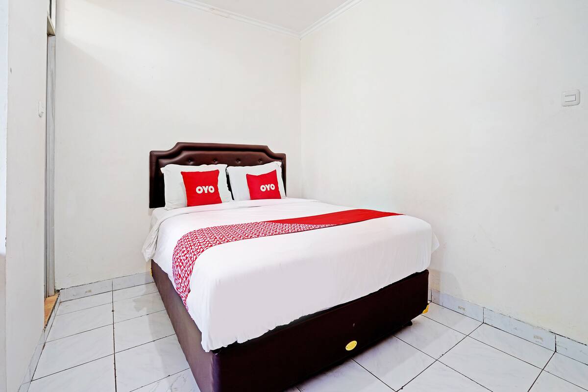 Un lit bien fait, recouvert d’une couvre-lit blanche et rouge, avec deux oreillers rouges ornés du mot « OYO », et une tête de lit en bois foncé, dans une pièce aux murs blancs et au sol carrelé.