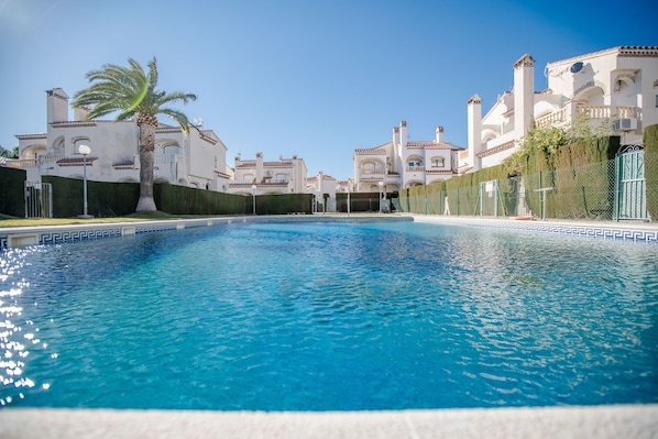 A swimming pool with a clear blue water surface, surrounded by white buildings with arched windows and balconies, and a palm tree in the foreground.