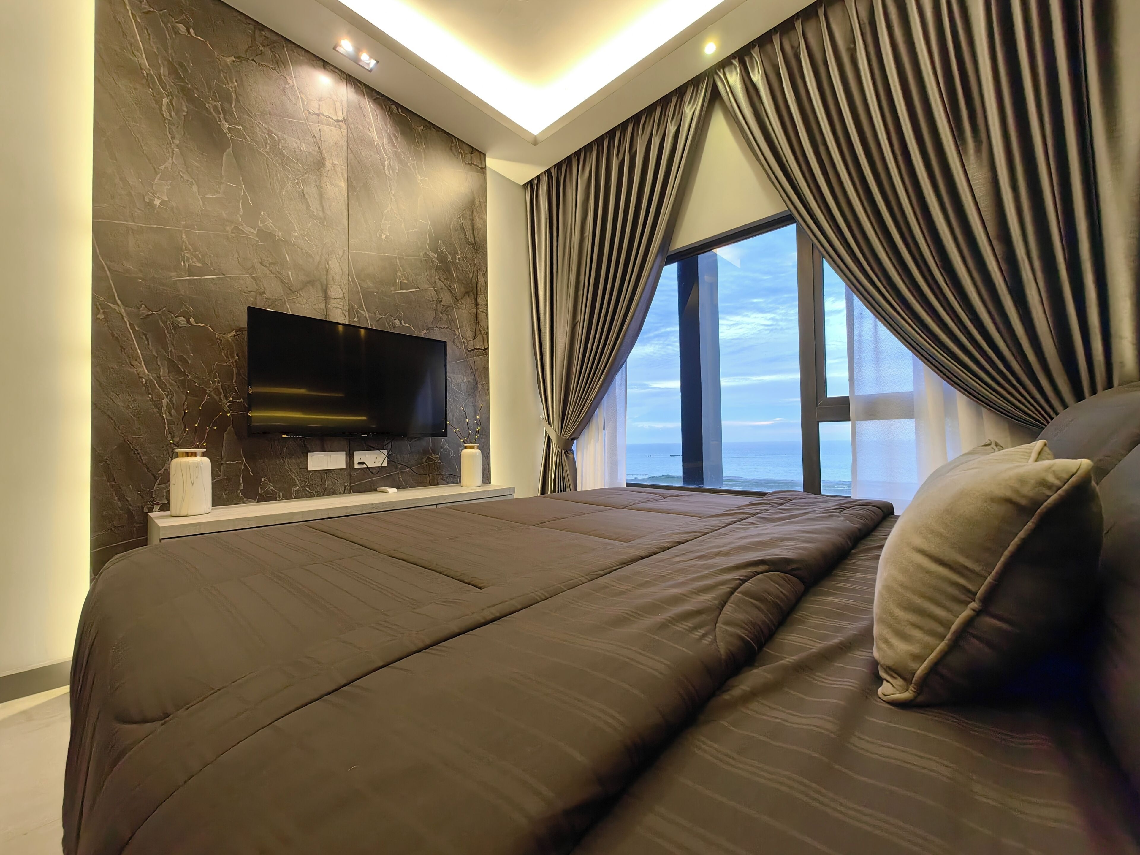 A modern bedroom with a large bed, a flat-screen TV mounted on a marble wall, and a view of the ocean through large windows.