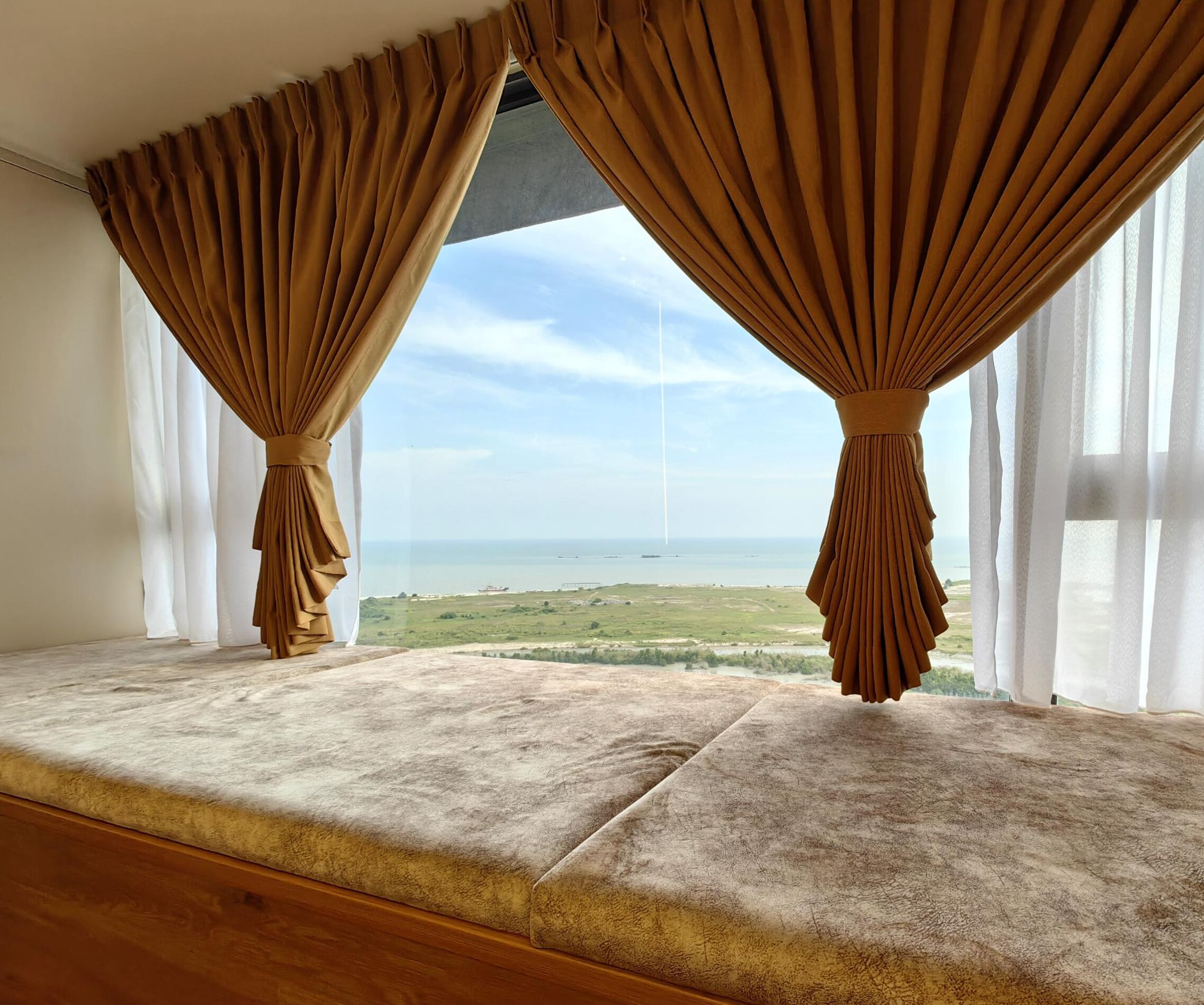 A room with a large window featuring brown curtains, a view of the sea, and a wooden bench.