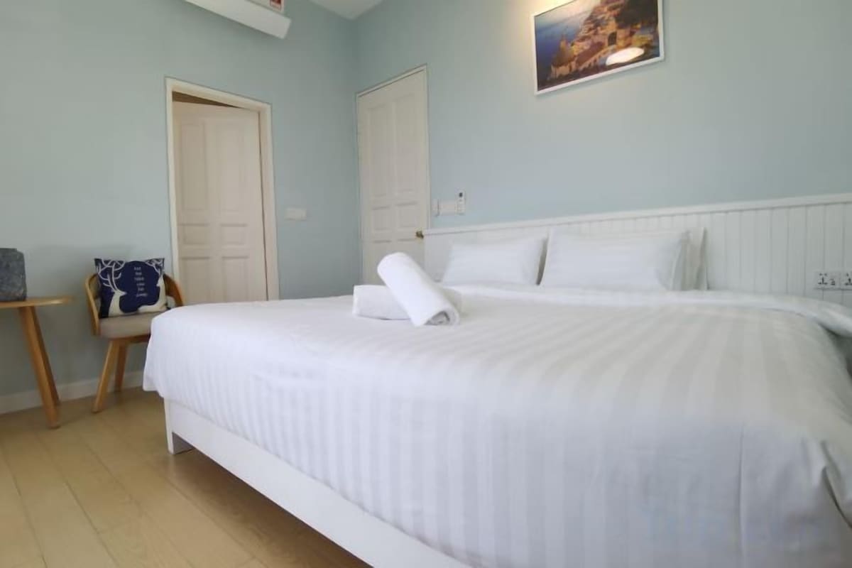 Un letto rifatto con lenzuola bianche e un asciugamano arrotolato, una sedia con un cuscino blu e una porta in una stanza dalle pareti azzurrine.