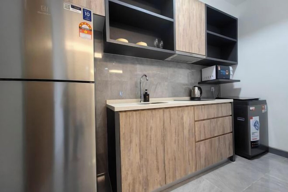 A modern kitchen with a stainless steel refrigerator, wooden cabinets, a countertop with a kettle, and a trash bin.