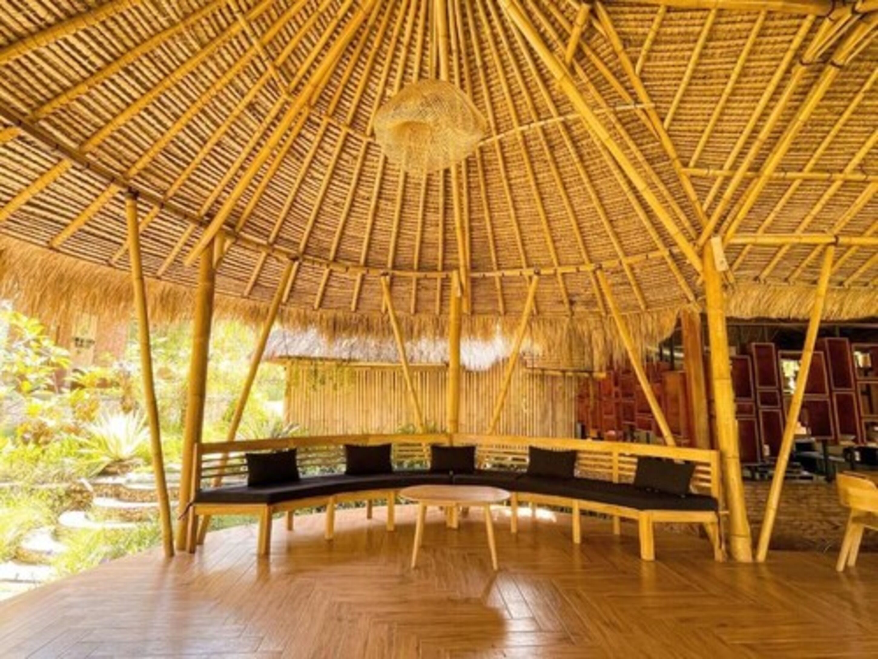 A thatched-roof structure with a curved bench and a round table, surrounded by wooden flooring and bamboo pillars.