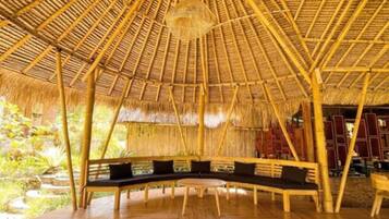 A thatched-roof structure with a curved bench and a round table, surrounded by wooden flooring and bamboo pillars.