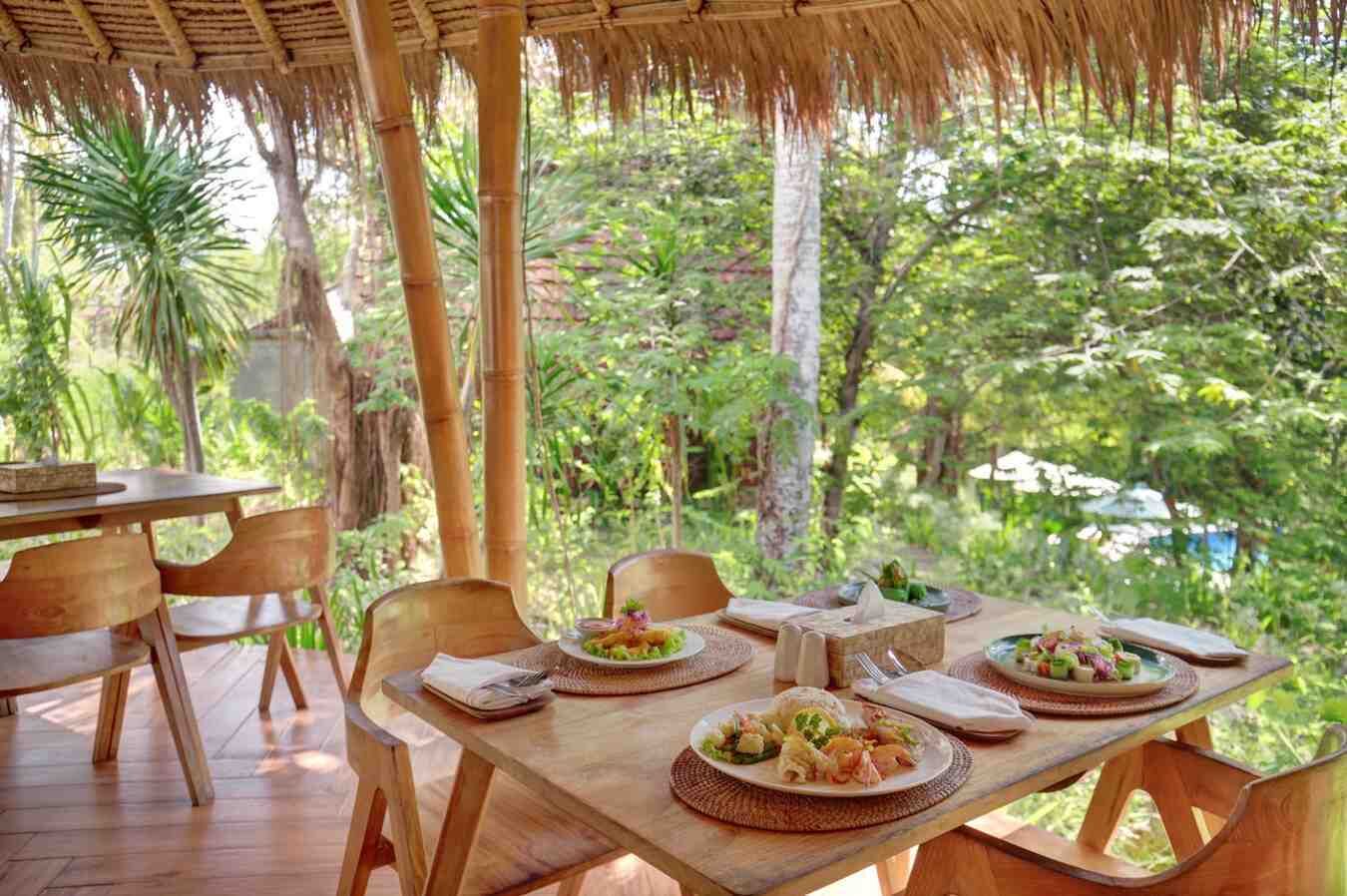 Un área de comedor al aire libre con mesas y sillas de madera, rodeada de exuberante follaje y estructuras de bambú.