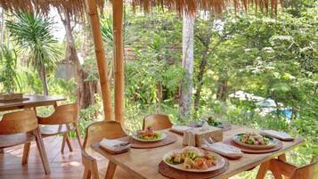 Un área de comedor al aire libre con mesas y sillas de madera, rodeada de exuberante follaje y estructuras de bambú.