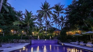 A pool area with palm trees, lounge chairs, and a stone wall. - Bila Penida Resort & Farm (Penida Island)