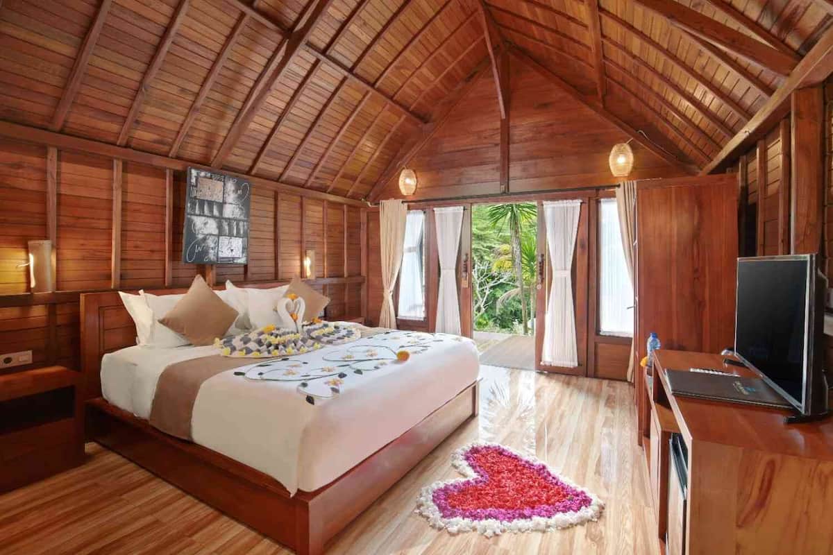 A wooden-themed bedroom with a bed, bedside tables, a desk with a TV, and a view of greenery outside.