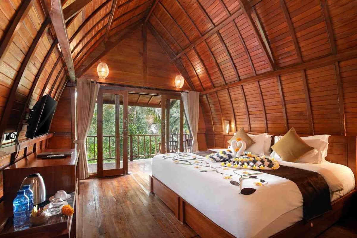A spacious, high-ceilinged room with a wooden ceiling and walls, a large bed with a heart-shaped pillow, a bedside table with a kettle, and a view of lush greenery outside.