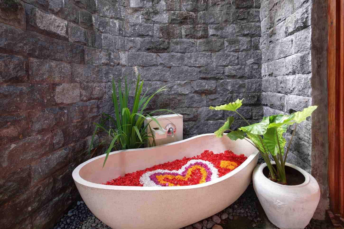 Une baignoire remplie de pétales de fleurs aux couleurs vives, entourée de plantes, le tout adossé à un mur de pierre.