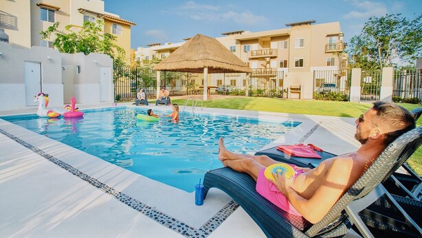 A swimming pool with lounge chairs, a thatched-roof structure, and modern buildings in the background.