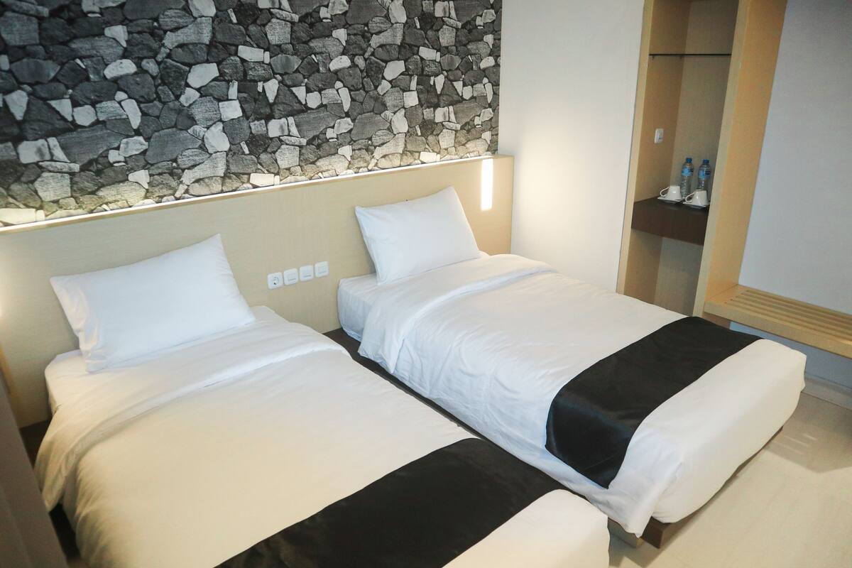 Ein Hotelzimmer mit zwei Betten, einer Steinsäule, einem kleinen Regal mit Wasserflaschen und einer Holzbank.