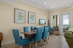 A dining area with a table and chairs, framed artwork on the wall, and a view of the ocean through a door.