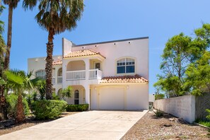 A two-story building with a balcony, a garage, and a well-maintained garden with palm trees and shrubs.